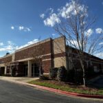 Modern office building with red brick and beige walls, large windows, and landscaped greenery.