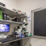 Lab desk with microscope, computer, and shelves of electronics and research supplies.